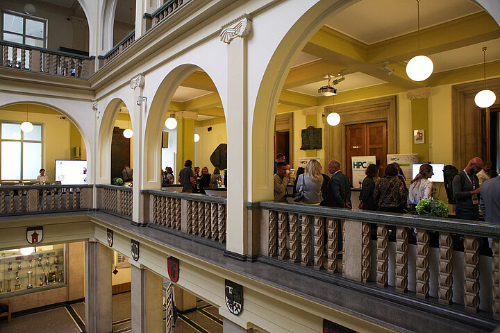 Na zdjęciu widać ujęcie na aulę gdzie stoją uczestnicy konferencji i rozmawiają, widać również fragment głównego holu budynku A-0