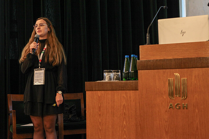 Na zdjęciu widać młodą studentkę przy mównicy wygłaszającą prezentację 