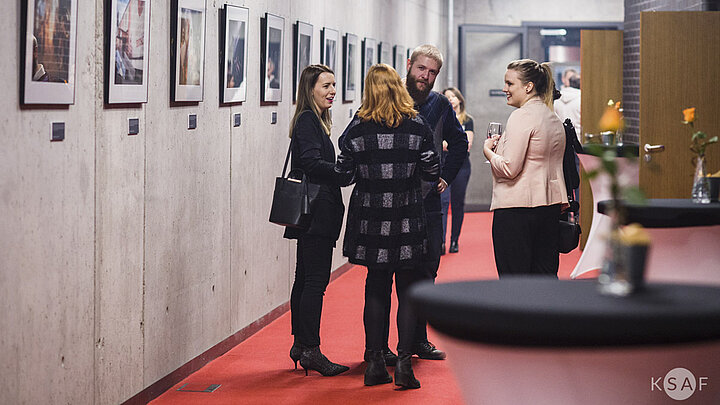 Czworo młodych ludzi w nowoczesnym, surowym holu. Na ścianach ekspozycja zdjęć.