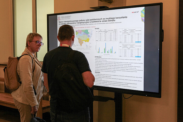 Na zdjęciu widać mężczyznę i kobietę stojących przed dużym monitorem, na którym wyświetlany jest poster