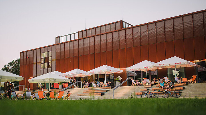 Nowoczesny, podłużny budynek o minimalistycznej elewacji wykonanej w rdzawej stalo kortenowej. Wyższa część jest ażurowa. Przed budynkiem na schodach siedzą młodzi ludzie.