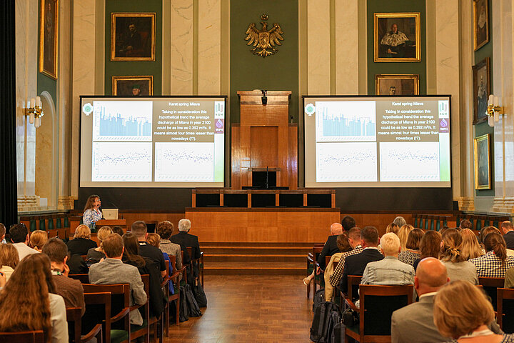 Na zdjęciu widać wnętrze auli wypełnione słuchaczami oraz rzut na ekrany wyświetlające prezentację